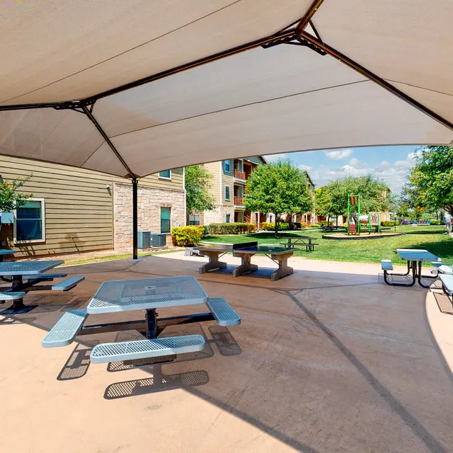 Outdoor Gathering Area Outdoor seating area with gray picnic tables under a large canopy, surrounded by greenery and buildings