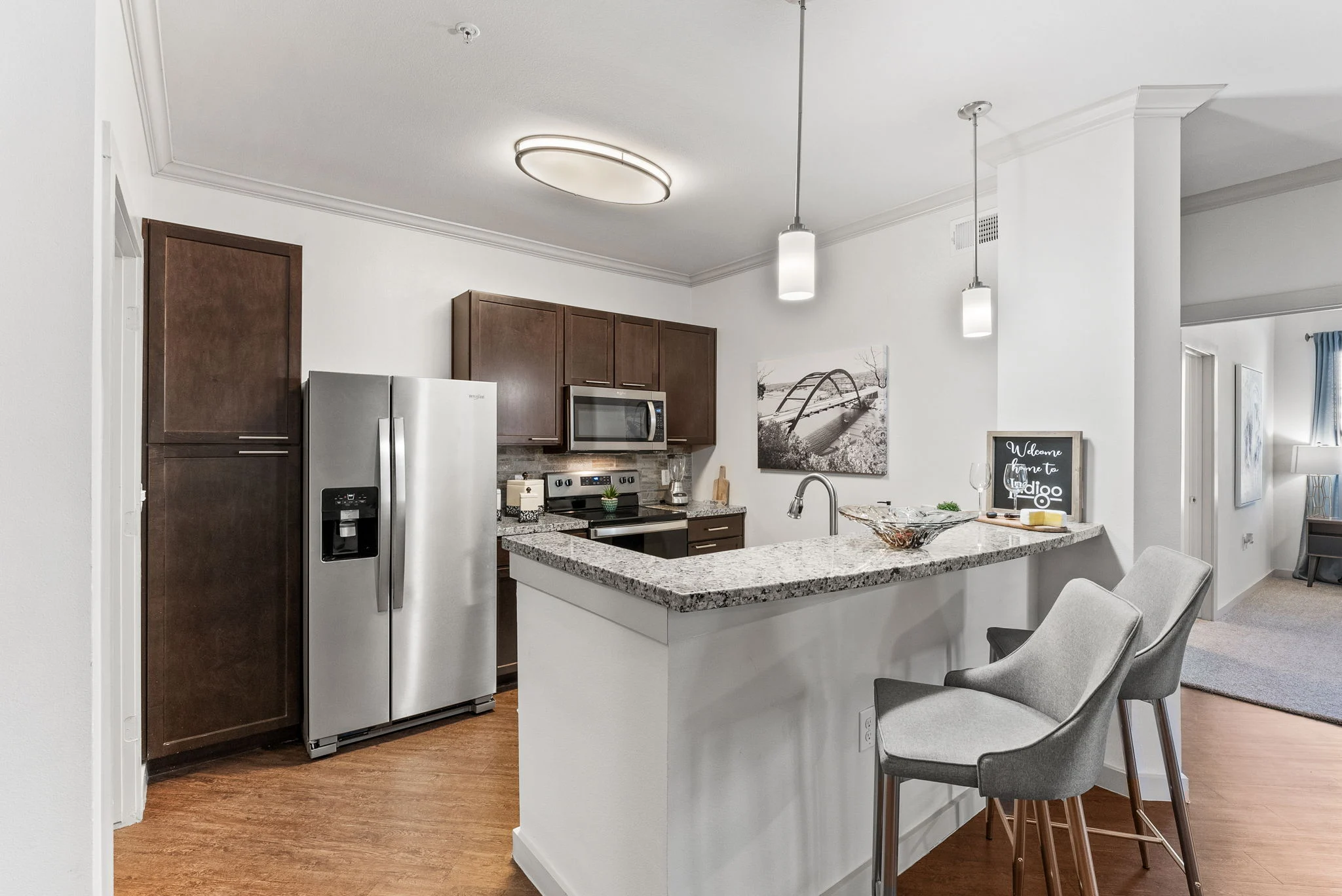 A modern kitchen with dark wooden cabinets and stainless steel appliances, including a refrigerator and microwave, along with a granite countertop and stylish bar stools. Natural light illuminates the space.