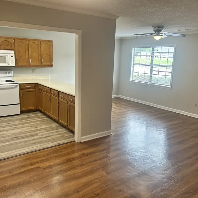 A spacious living room connected to a kitchen, featuring wooden cabinets, white appliances, and a window letting in natural light.