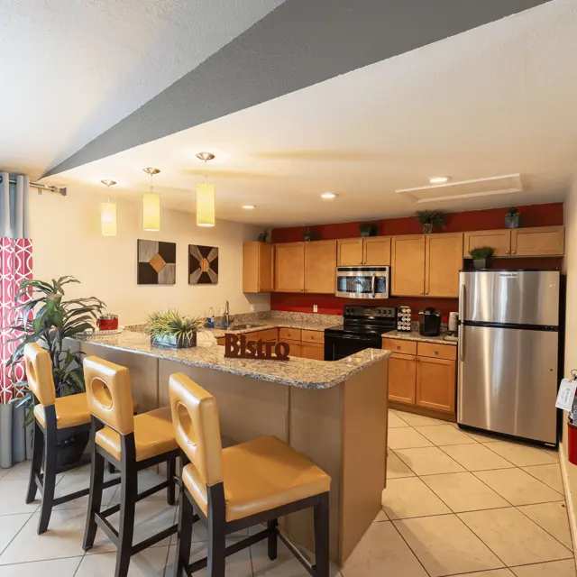 A modern kitchen featuring wooden cabinets, stainless steel appliances, and a large island with seating. The space is bright with natural light filtering through a window, complemented by decorative plants and colorful curtains.