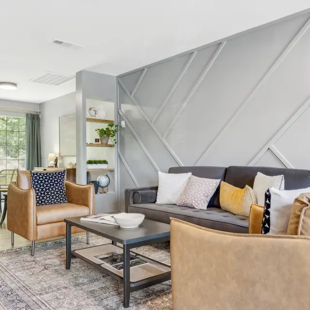 Modern living room with a gray geometric accent wall, cozy sofa, armchairs, and a dining area visible in the background.