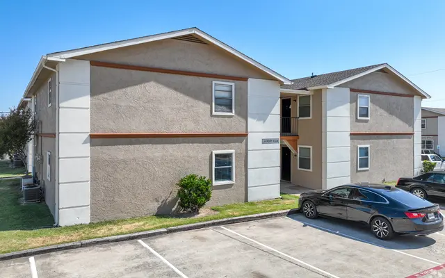 A picture of a two-story apartment complex with beige and white exterior, showing two sides of the building, parking spaces in front, and several windows.
