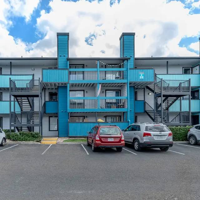 Exterior view of a modern apartment complex with blue accents and multiple parking spaces in front.