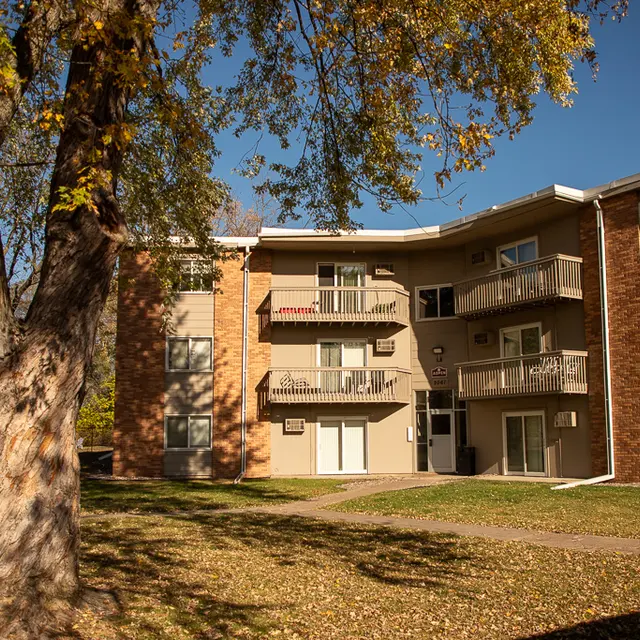 A multi-story apartment building with brick and siding exterior, surrounded by green grass and a large tree in the foreground.