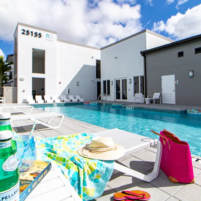 Inviting Swimming Pool Area A sunny swimming pool area featuring lounge chairs, a sandy blue towel, and a bottle of sparkling water in the foreground. The pool is clear with palm trees in the background, and modern white buildings line the poolside.