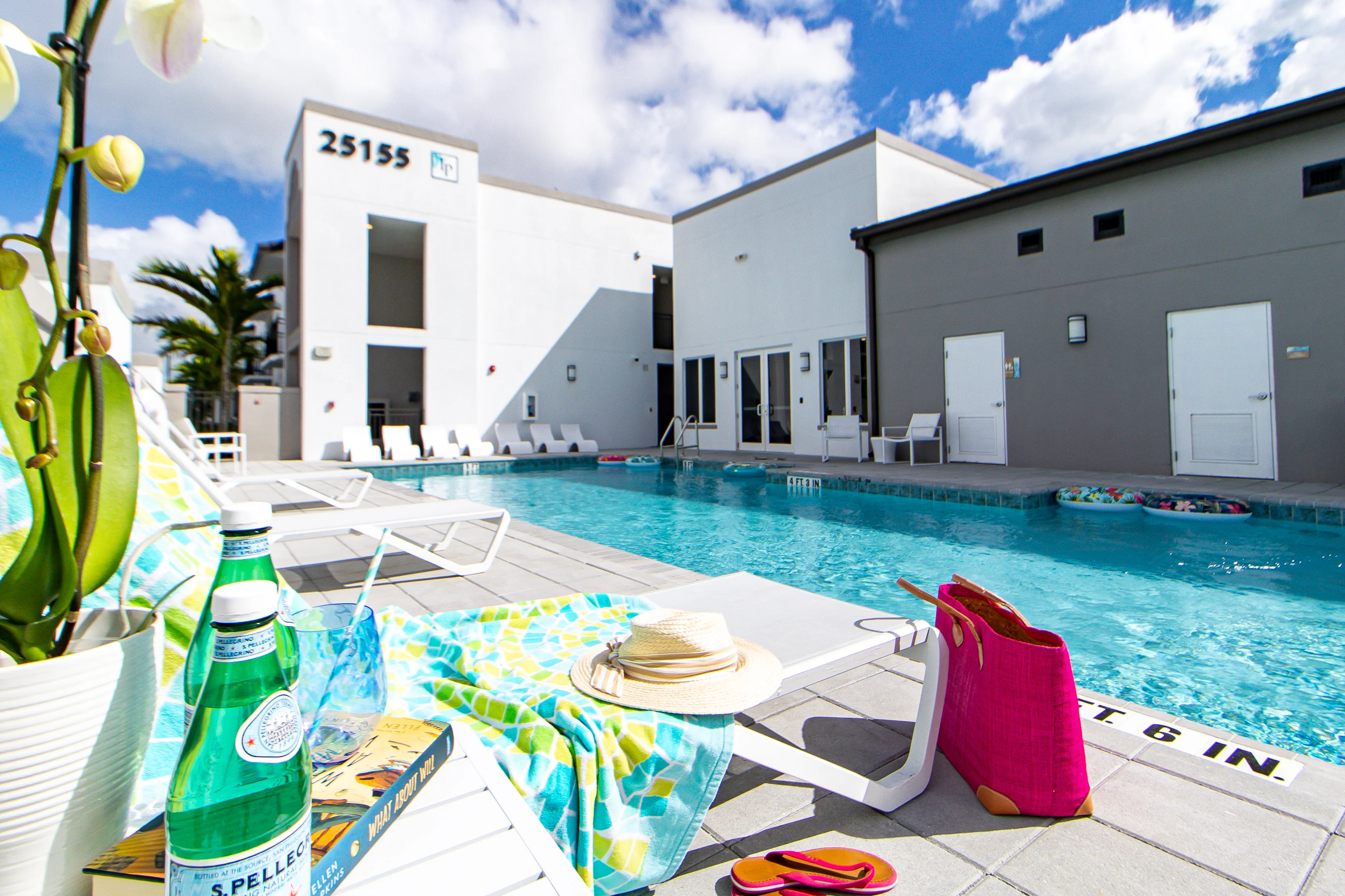 Inviting Swimming Pool Area A sunny swimming pool area featuring lounge chairs, a sandy blue towel, and a bottle of sparkling water in the foreground. The pool is clear with palm trees in the background, and modern white buildings line the poolside.