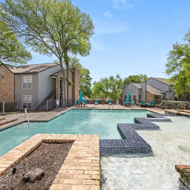 A sunny pool area with modern landscaping featuring two separate sections of water, surrounded by brick walkways and green trees. There are lounge chairs along the poolside.