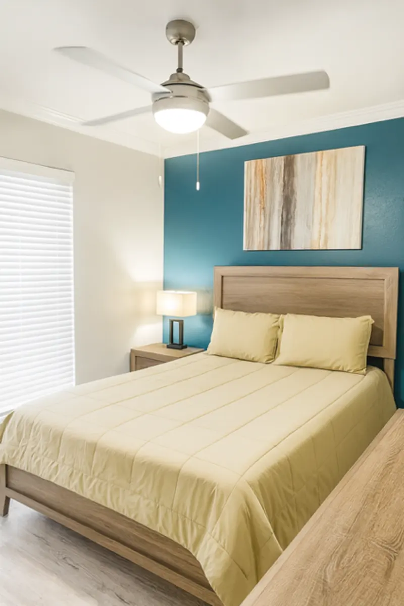 Modern Bedroom Interior A modern bedroom featuring a queen-sized bed with a beige comforter, a wooden bed frame, and a matching nightstand. There is a lamp on the nightstand, and a blue accent wall with abstract artwork. A ceiling fan is overhead, and a window with blinds is visible.