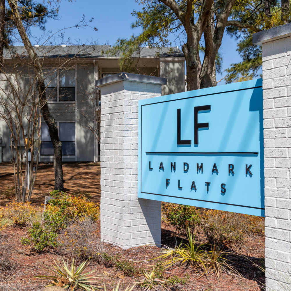 A light blue sign displays 'LF' and 'Landmark Flats' in white lettering. It is positioned in front of a residential building surrounded by trees and grass.