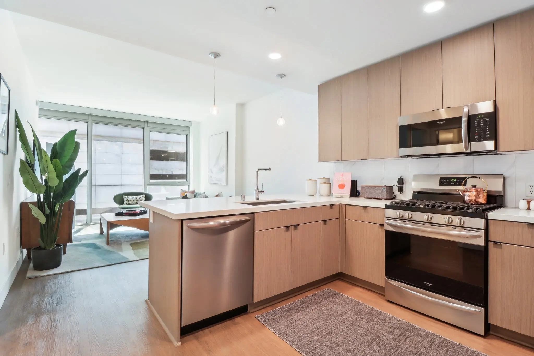 A modern kitchen with light wood cabinetry, stainless steel appliances, and an open layout leading to a living area with natural light.