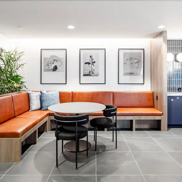 A modern lounge area featuring a curved brown upholstered bench, black chairs, a round table, and framed art on the wall. There is a plant in the corner and a kitchenette with blue cabinetry in the background.