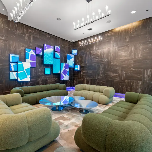 A modern lounge area featuring green rounded sofas arranged around a glass coffee table, with a wall displaying colorful artwork and subtle lighting.