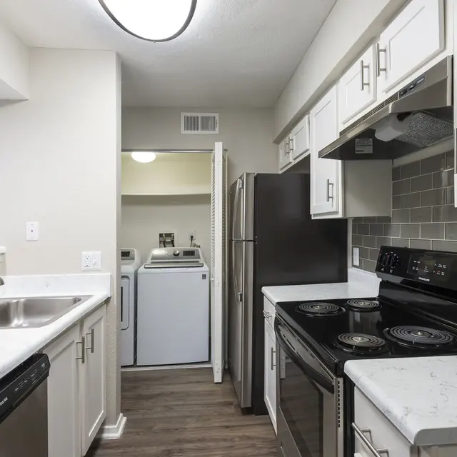 Modern kitchen with white cabinets, stainless steel appliances, and a laundry area visible in the background.