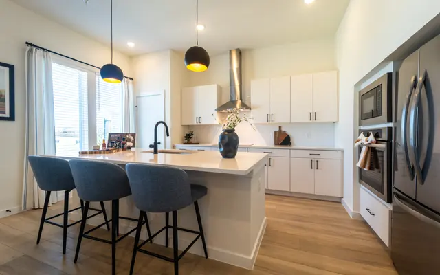A modern kitchen with a large island, three bar stools, white cabinetry, and stainless steel appliances. There are pendant lights hanging above the island and a window with light curtains.