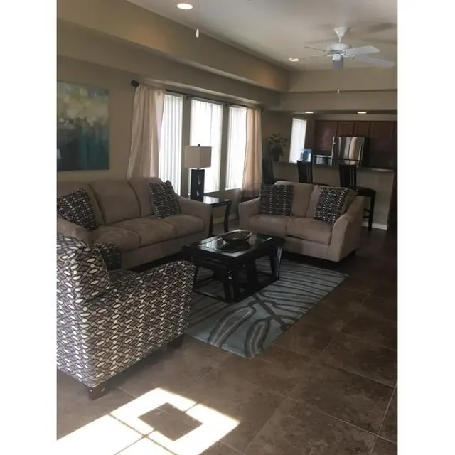 A cozy living room featuring a beige sofa set, a patterned accent chair, and a coffee table on a geometric rug. Natural light comes through large windows with sheer curtains, and kitchen appliances are visible in the background.