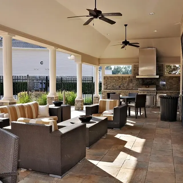 An outdoor lounge area featuring comfortable seating, a stone grill area, and a fenced pool in the background.