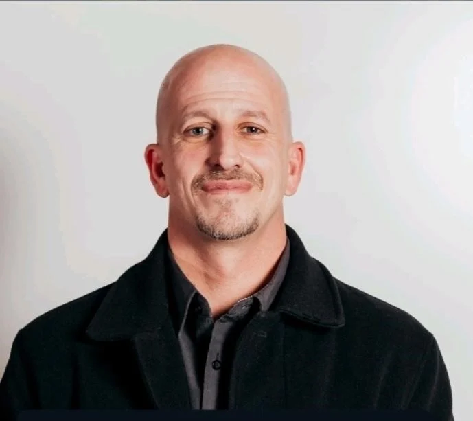 Portrait of a Man in a Black Coat A man with a shaved head, wearing a black coat over a dark shirt, smiling at the camera against a plain background.
