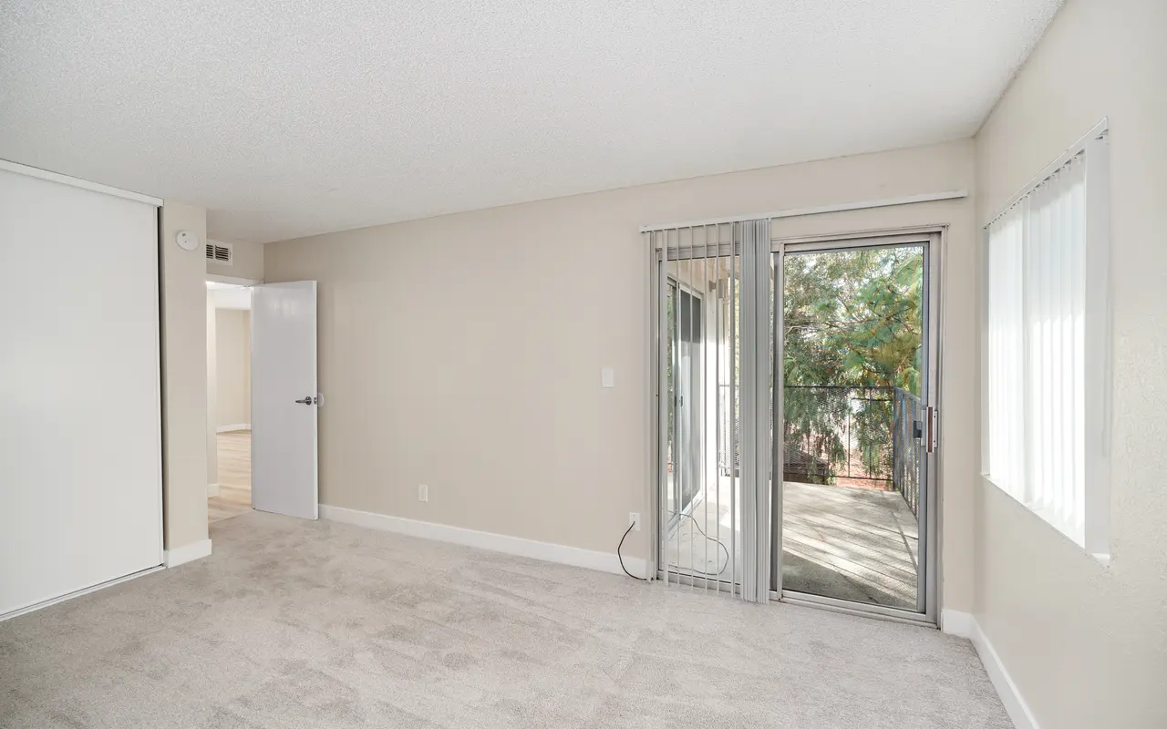 A spacious empty living room with light-colored carpet, featuring a sliding glass door leading to a small balcony. The walls are painted a soft neutral shade, and there are two vertical blinds windows letting in natural light.