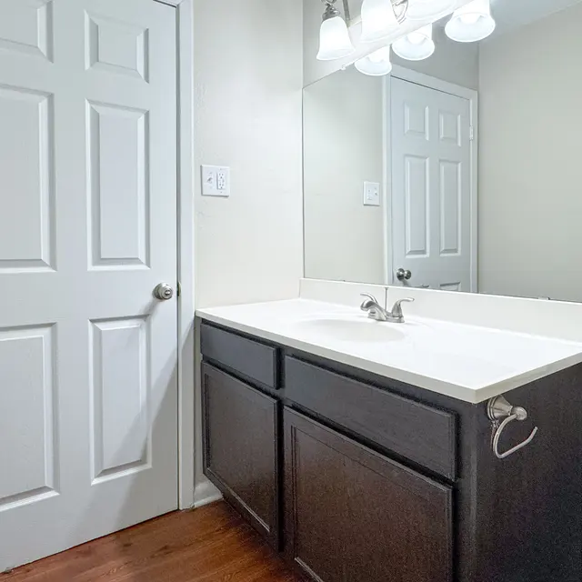 A clean and simple bathroom featuring a white countertop, dark cabinets, and a large mirror. The room has a light-colored wall and wooden flooring with a door on the left side.