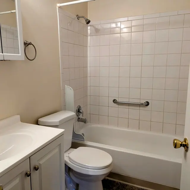 A clean and simple bathroom featuring a white bathtub with a shower, a toilet, and a sink with a cabinet. The wall tiles are white and the decor is neutral.