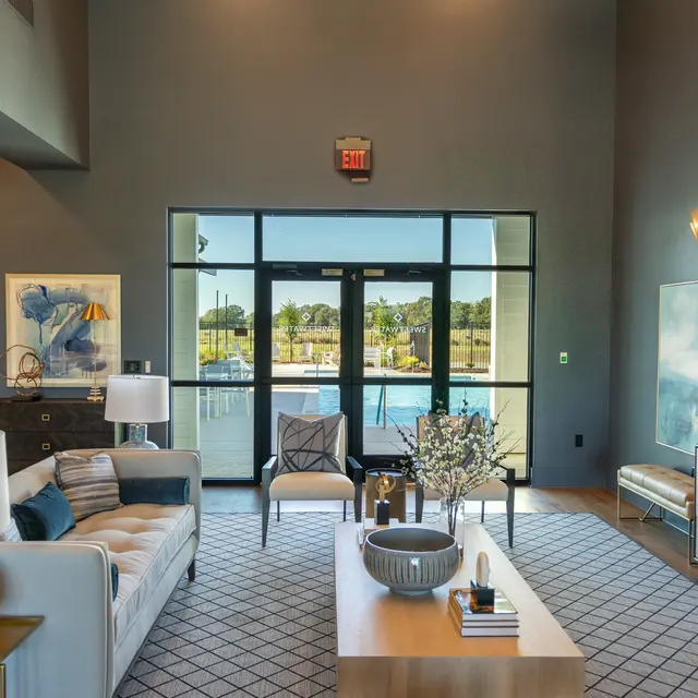 A modern living room with large windows overlooking a patio area. The space features a beige sofa, stylish chairs, a decorative coffee table, and various artworks on the walls. Soft lighting and a contemporary design are evident, creating a cozy atmosphere.
