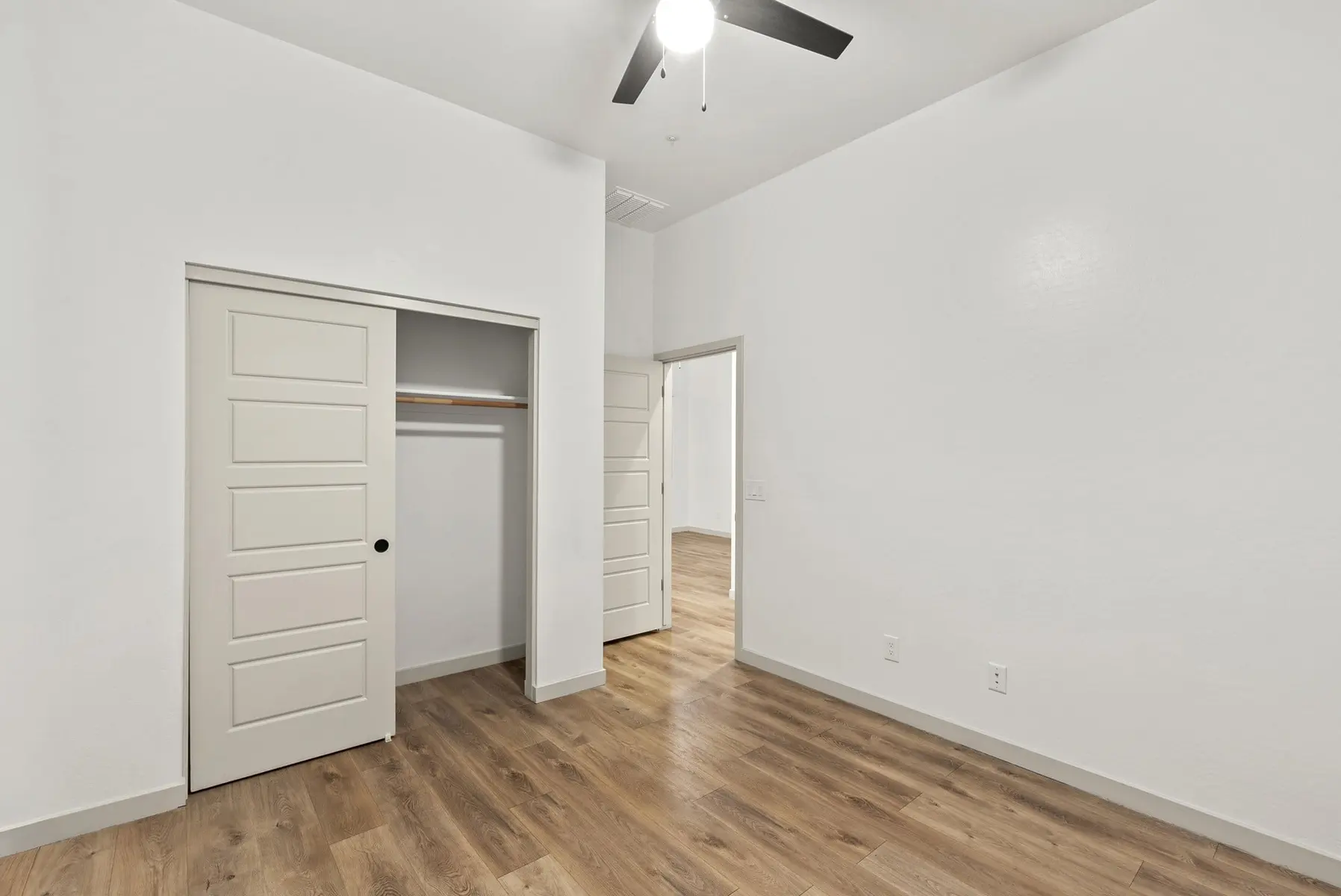 A well-lit, minimalistic room with a ceiling fan, open closet, and light-colored walls and flooring.