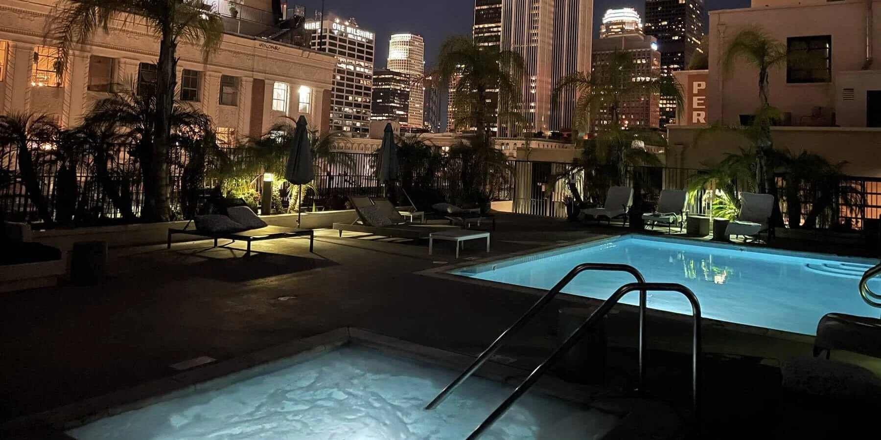 A rooftop pool area at night with city skyscrapers in the background, featuring a lit pool, a hot tub, lounge chairs, and palm trees.
