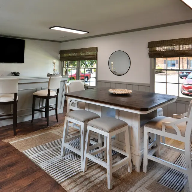 A modern dining area featuring a rectangular table with white chairs, a bar with high stools, and a large window with blinds. There is a flat-screen TV mounted on the wall and a decorative mirror.