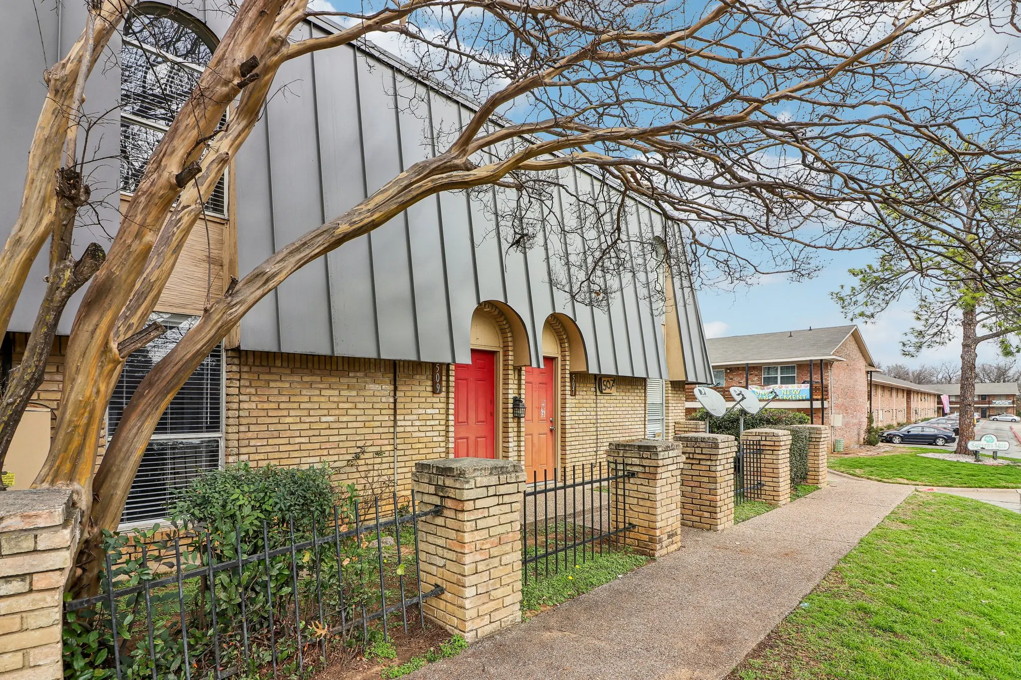 Exterior view of a modern apartment building with brick walls and unique metal roof design. Two red doors are visible at the entrance, with a wrought iron fence and landscaped garden.