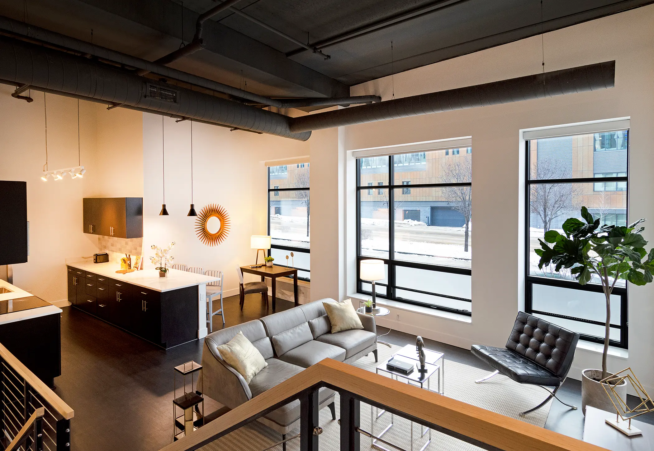 A spacious modern loft apartment featuring a sleek grey sofa, large windows with a view, an open kitchen with stylish fixtures, and contemporary furniture.