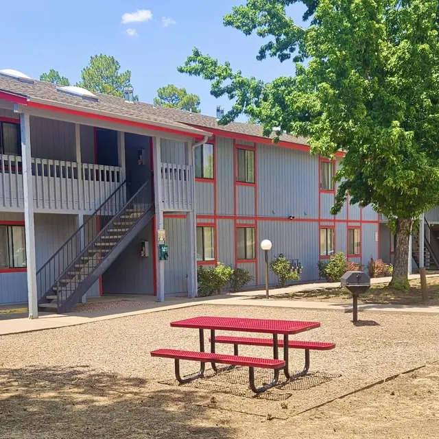 Apartment Complex Outdoor Area An outdoor view of an apartment complex featuring two-story buildings with red accents. A central area includes a red picnic table and a grill, surrounded by trees and landscape.