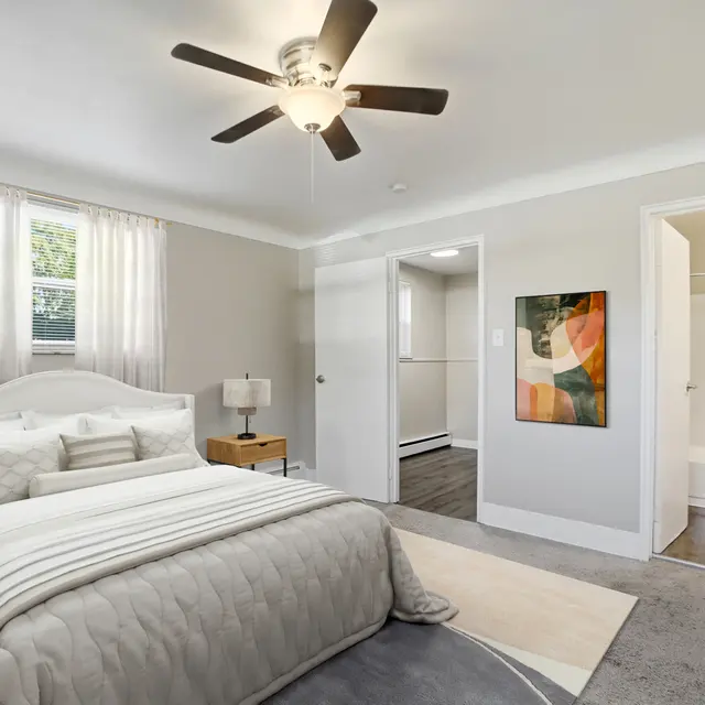 A modern bedroom with a large bed, a ceiling fan, and a white rug on the floor. There are two windows with sheer curtains and a painting on the wall. A bathroom door is visible on the right.