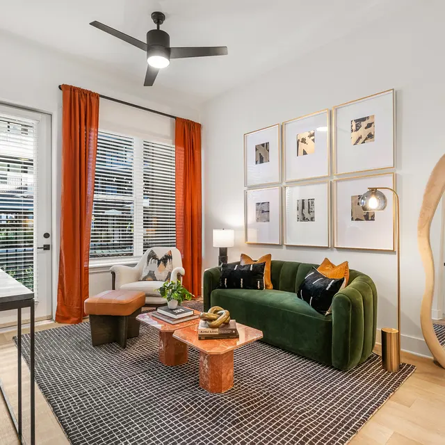 A modern living room featuring a green velvet sofa with orange and black decorative pillows, a wooden coffee table, and a mirror. The room is well-lit with large windows covered by sheer curtains and has a textured area rug.