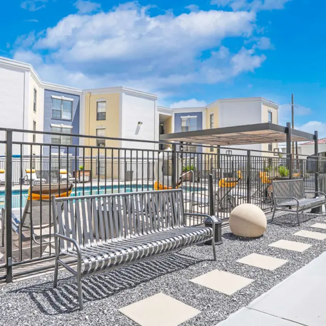 A sunny poolside area featuring modern benches, decorative stone pathways, and a swimming pool surrounded by buildings.