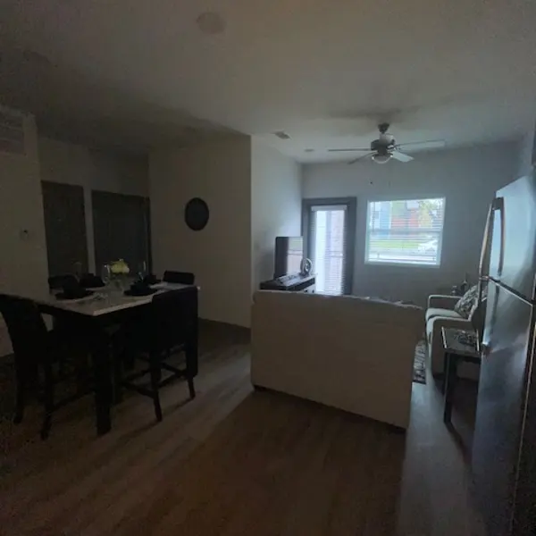 A dimly lit living space featuring a small dining area with chairs, a sofa facing a TV, and a ceiling fan. A window allows some natural light to enter from the right side of the image.