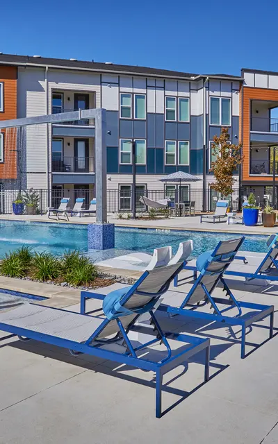 A modern apartment complex with a swimming pool, lounge chairs, and landscaped areas. The pool is surrounded by patio furniture and greenery, with several apartment buildings in the background.