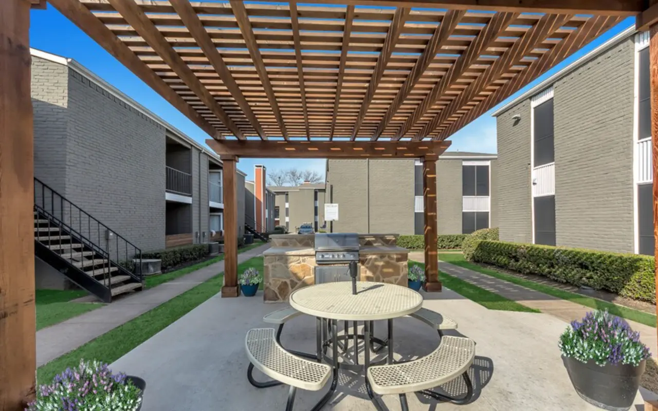 An outdoor pergola with a picnic area underneath, featuring a stone grill and planters, surrounded by apartment buildings.
