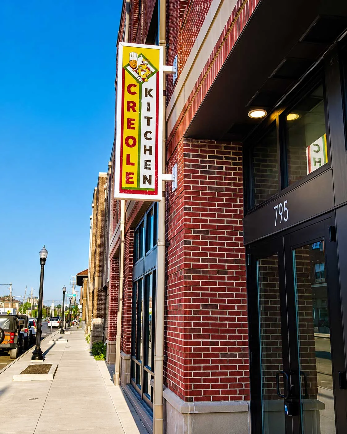 A brightly colored sign for 'Creole Kitchen' hangs above a brick building, with a clear blue sky in the background. The street and sidewalk are visible, along with a lamppost on the left.