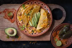 Tacos, beans, cheese, chills, and carrots presented on a plate. Plate resting on a wooden chopping board with avocado and seasonings spilled across it. black table.