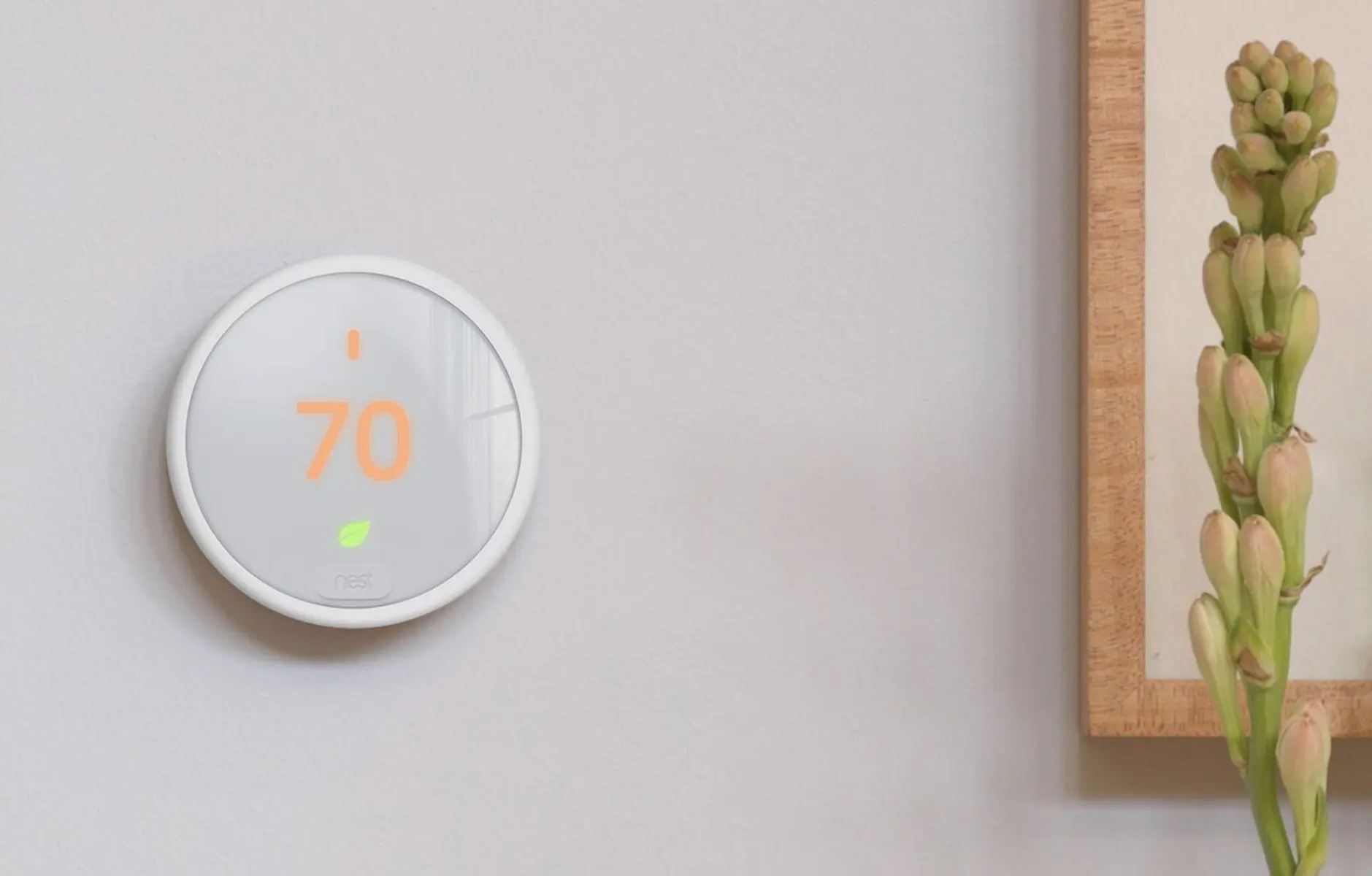 A modern smart thermostat displaying a temperature of 70°F on a light grey wall, next to a plant