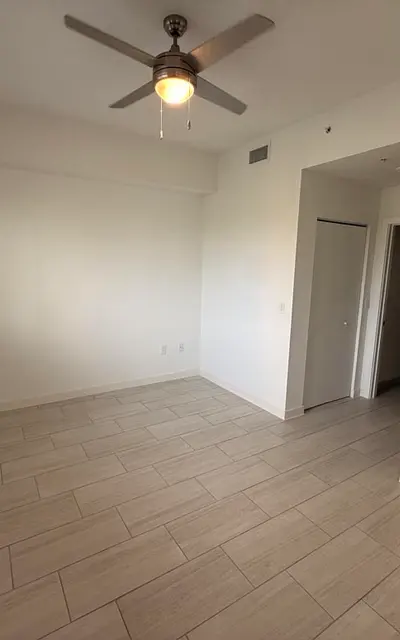 A spacious, empty room with light-colored tiled flooring, a ceiling fan, and a window with blinds letting in natural light. There is a door leading to another room on the right side.