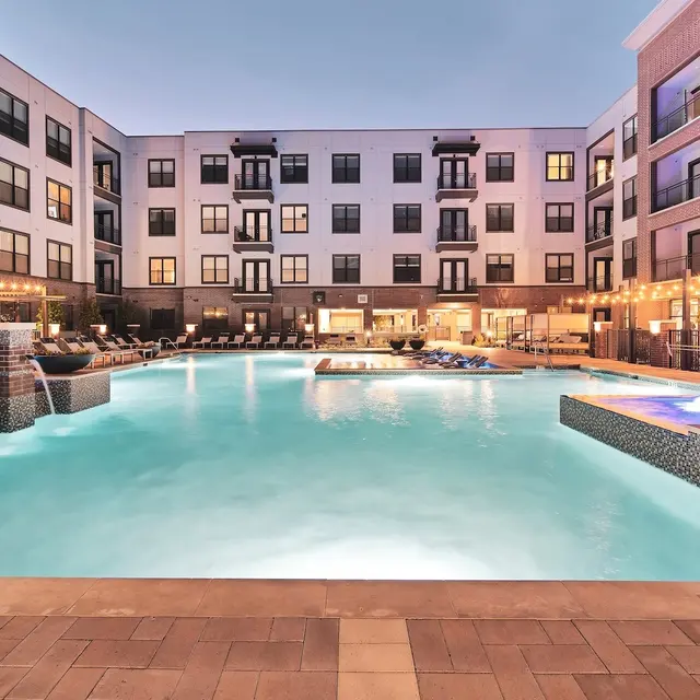 Luxurious Apartment Pool Area A luxurious apartment complex featuring a large swimming pool surrounded by contemporary buildings. The pool has waterfalls and a hot tub, with lounge chairs placed around it and warm lighting creating an inviting atmosphere at dusk.