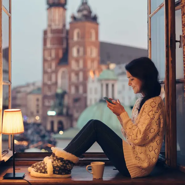 Woman sitting on a windowsill, wearing cozy clothes, using a phone with a cityscape view in the background at dusk.