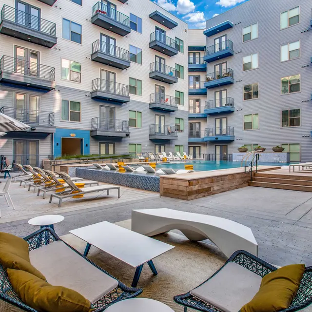 A modern apartment complex outdoor pool area, featuring lounge chairs, umbrellas, and seating arrangements. The pool is surrounded by contemporary architecture with multiple levels.