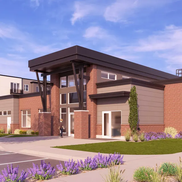 A modern building exterior featuring a mix of brick and metal construction, with a large entrance and landscaped areas including purple flowers. Nearby, there are additional buildings and people walking.