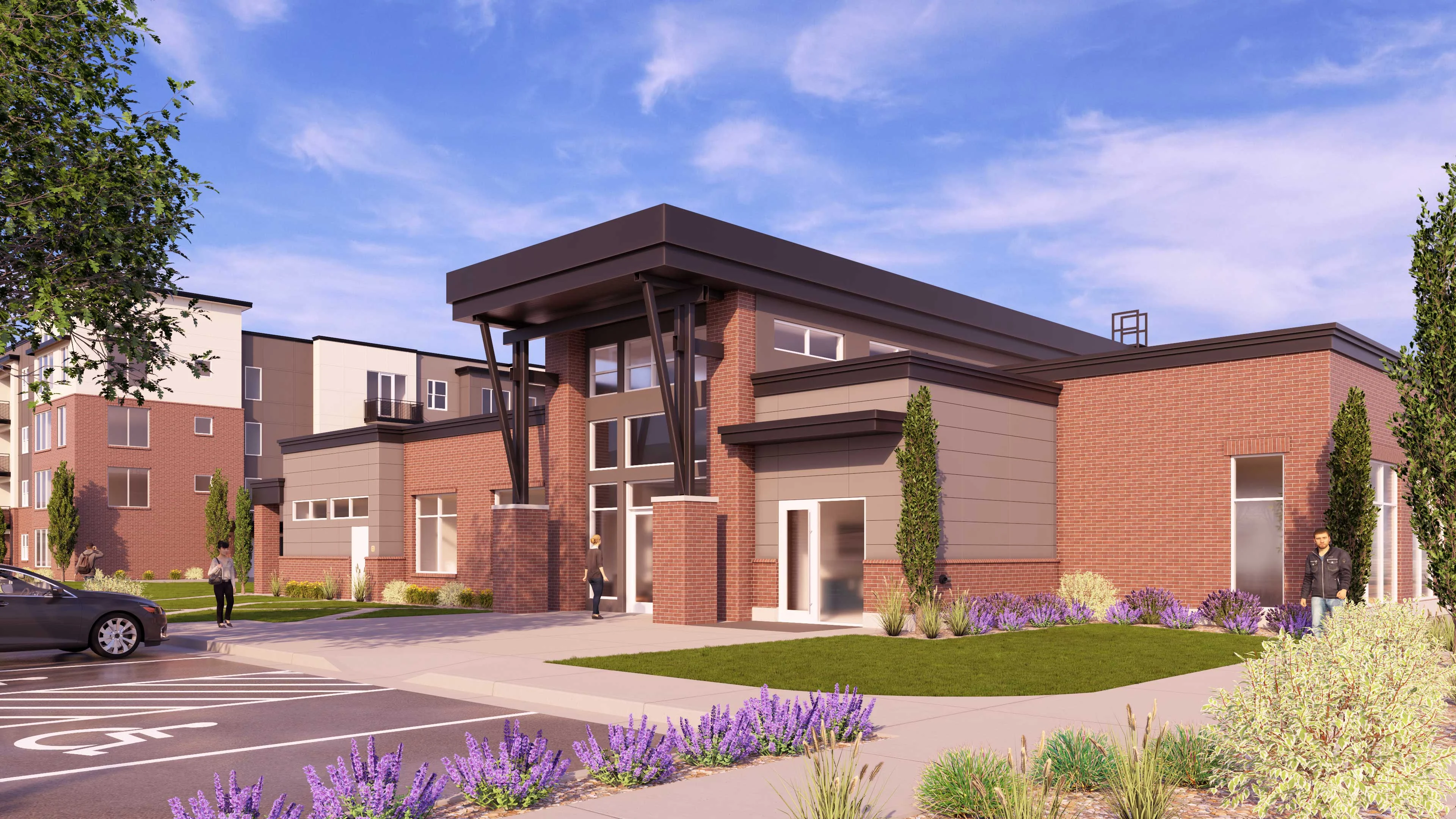 A modern building exterior featuring a mix of brick and metal construction, with a large entrance and landscaped areas including purple flowers. Nearby, there are additional buildings and people walking.