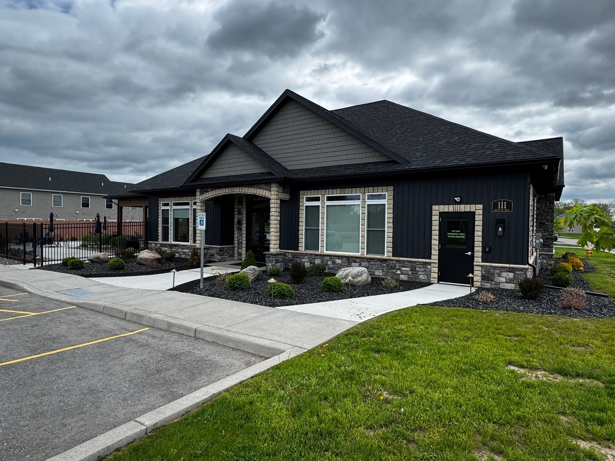 A modern single-story building with a dark exterior and stone accents, set against a cloudy sky. The building features multiple windows and a neatly landscaped area in front. A parking lot is visible in the foreground with marked parking spaces.