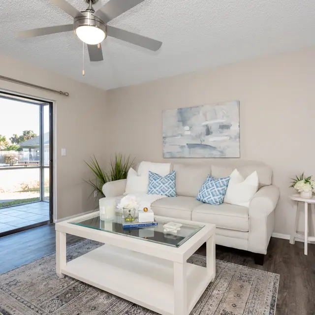 A modern living room featuring a light sofa with blue and white cushions, a glass coffee table, and a decorative rug. There are large windows with curtains, offering a view of an outdoor space. The room has a neutral color scheme and plants for decoration.