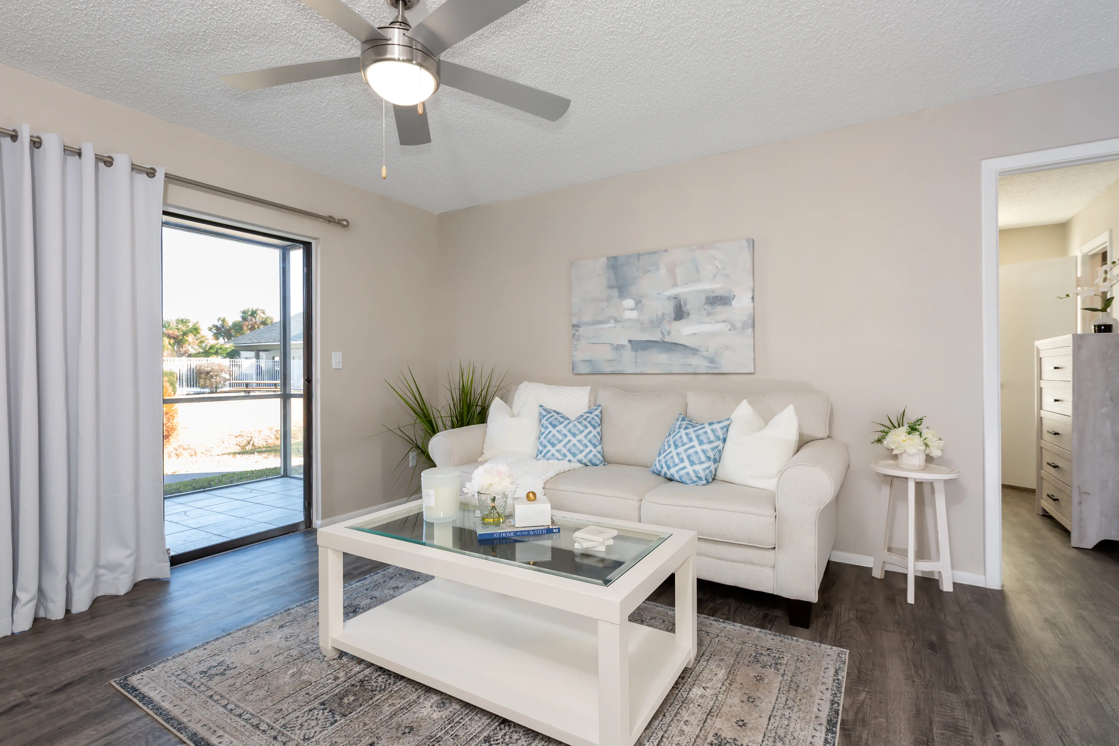 A modern living room featuring a light sofa with blue and white cushions, a glass coffee table, and a decorative rug. There are large windows with curtains, offering a view of an outdoor space. The room has a neutral color scheme and plants for decoration.