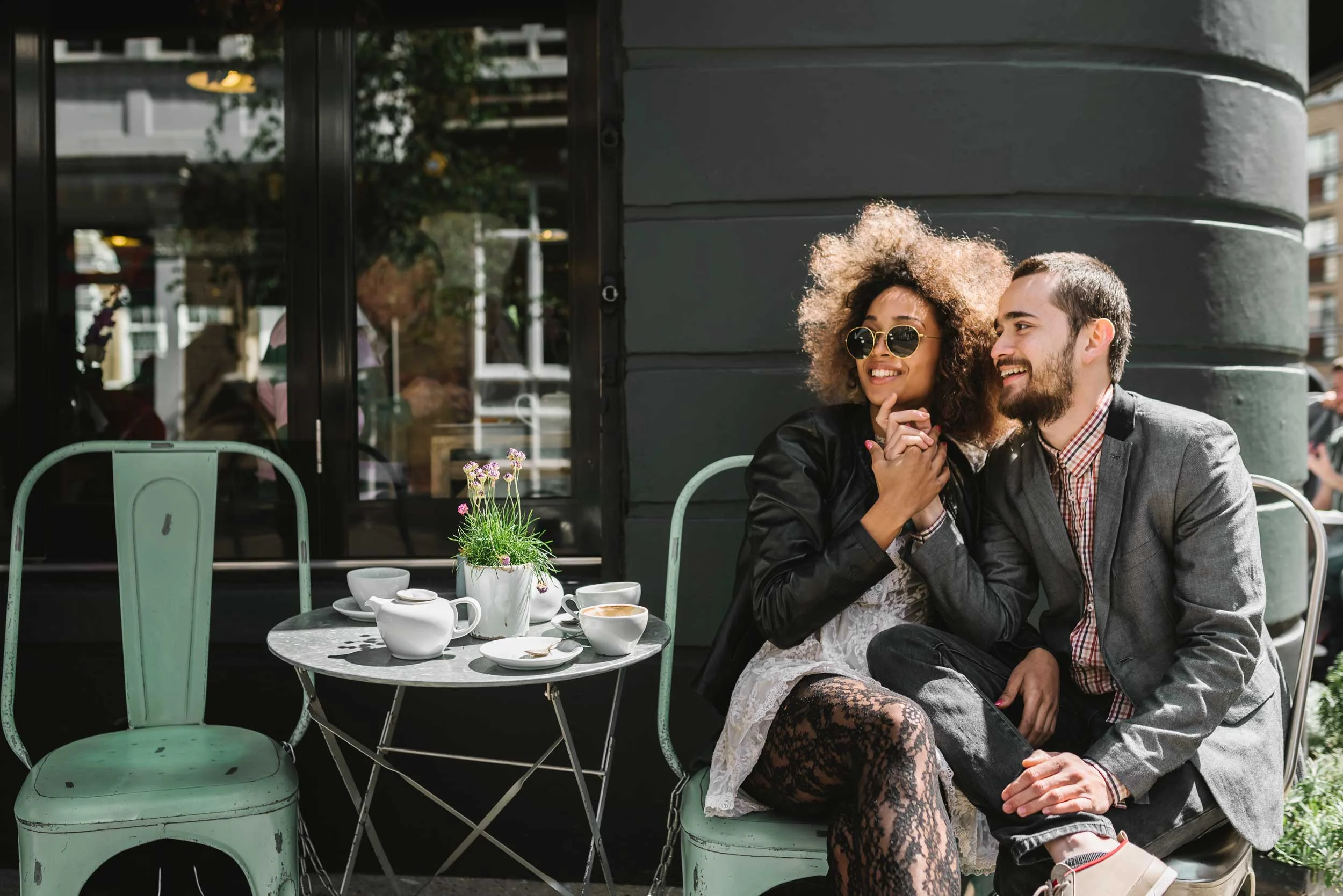 Couple at Cafe A stylish couple sitting outside a cafe, smiling and enjoying each other's company. The woman has curly hair and sunglasses, wearing a black jacket and patterned tights. The man, with a beard, is dressed in a gray blazer and shirt. A small table with tea or coffee cups is in front of them.